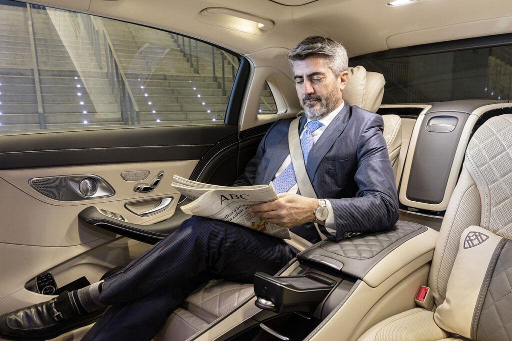 Man in suit reading newspaper in a luxury car's backseat.