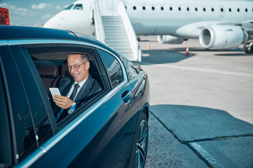 Businessman checking phone inside luxury car near private jet.
