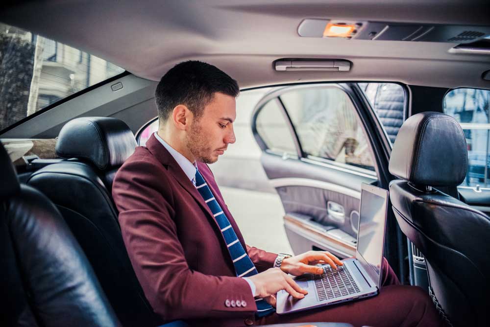A man in a suit working on a laptop inside a car.